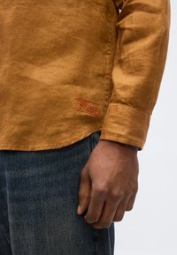 Καφέ πουκάμισο με μακριά μανίκια, ορατό μανσέτα και λεπτομέρεια κόκκινης ραφής κοντά στη ραφή, φορεμένο με μπλε τζιν, χέρι χαλαρό στο πλάι.