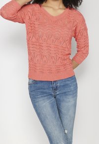 Pull en maille corail avec col en V, manches trois-quarts et motif texturé. Associé à un jean en denim bleu.