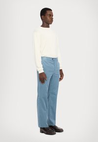 Lichtgekleurde gebreide trui, blauwe corduroy broek en bruine schoenen. Model staat zijwaarts tegen een effen achtergrond en laat de details van de outfit zien.