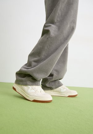 Białe sneakersy z teksturowanym wierzchem, sznurowanym designem i gumową podeszwą. Noszone z szarymi, prążkowanymi spodniami w stylu wide-leg na zielonej powierzchni.