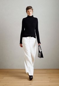 Pull-over noir à col roulé côtelé, pantalon large blanc et sac à main noir. Le mannequin marche sur un sol en bois contre un mur gris clair.