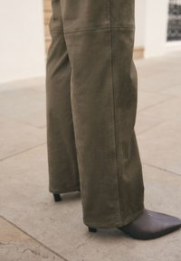 Pantalon large couleur vert olive en tissu doux, avec des détails de coutures, porté avec des chaussures pointues marron foncé.