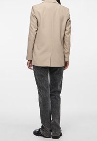 Blazer beige de corte recto, con una abertura sencilla en la parte posterior, combinado con pantalones grises texturizados y zapatos negros gruesos.