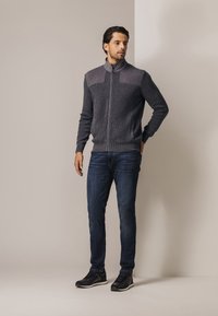 Grijze zip-up trui met een textuur op het bovenstuk, geribbelde zoom, gecombineerd met een donkere spijkerbroek en zwarte sneakers. Eenvoudig, modern ontwerp.