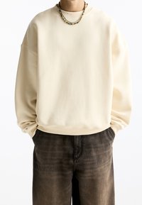 Sweter w kolorze kremowym o luźnym kroju, z prążkowanymi mankietami i miękką teksturą, w połączeniu z ciemnymi, wyblakłymi spodniami. Casualowy design.