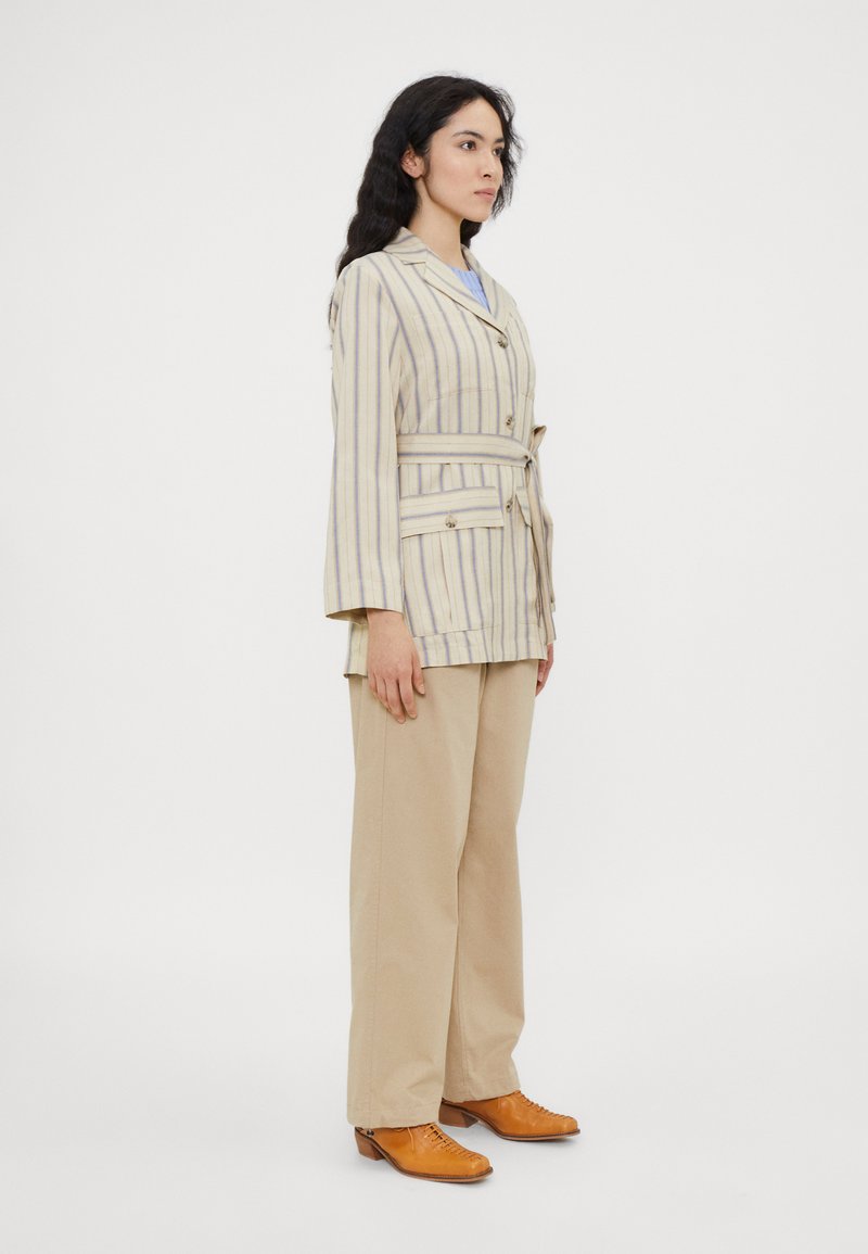 Beiger gestreifter Blazer mit Gürtel, weiten Ärmeln und vorderen Knöpfen. Kombiniert mit beigen Hosen und braunen Lederschuhen. Schlichtes und strukturiertes Design.