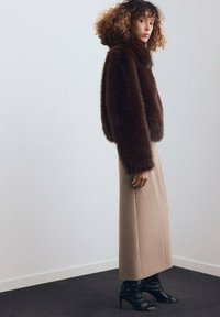 Chaqueta de pelaje sintético marrón, corta con un cuello alto. Falda larga de corte ajustado beige y botas negras de tacón. Diseño simple y moderno sobre un fondo claro.