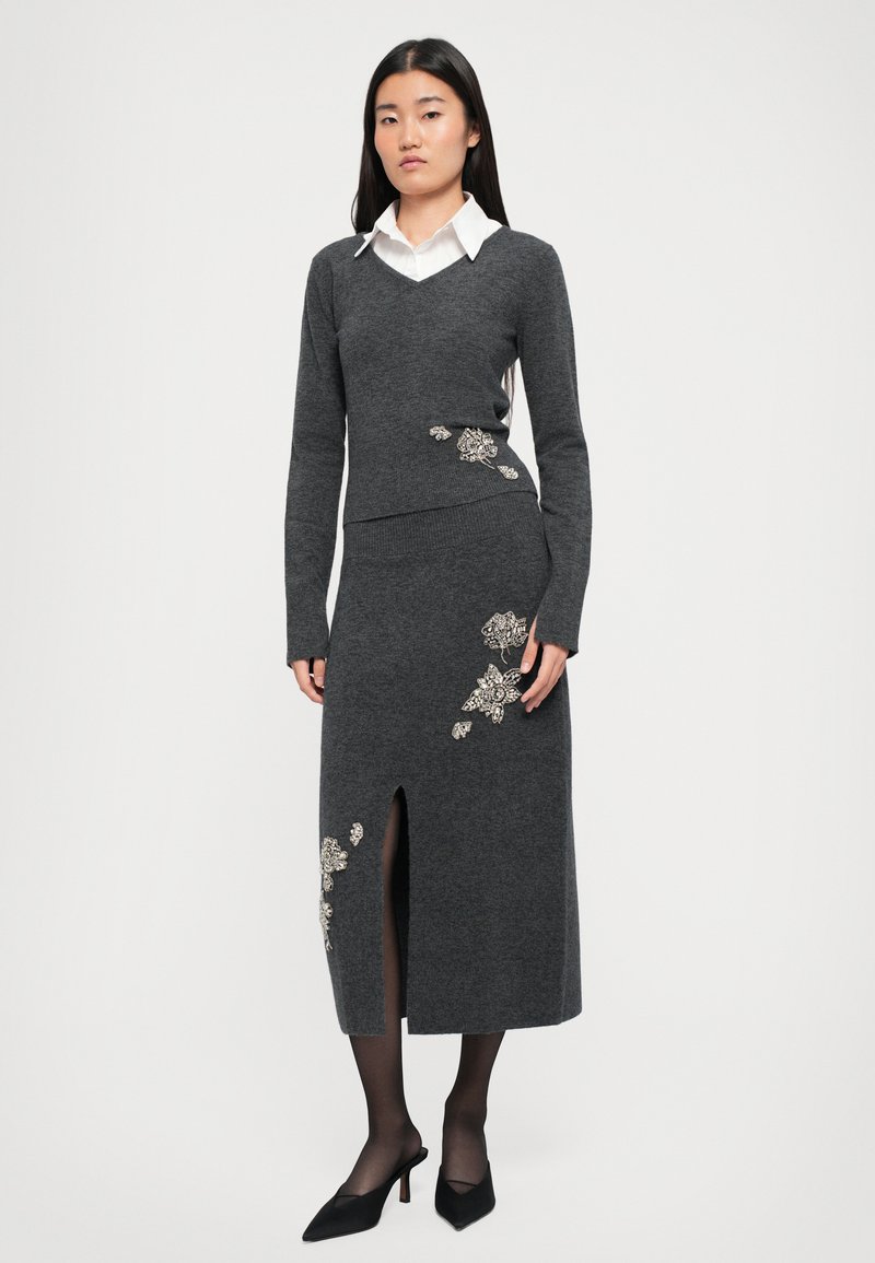 Frau trägt einen dunkelgrauen Strickpullover und einen passenden Midirock mit floraler Stickerei und einem vorderen Schlitz, kombiniert mit schwarzen Heels und Strumpfhosen.