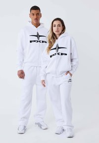 Sudaderas blancas con cremallera y logo negro "PXP", combinadas con pantalones de chándal a juego. Ambos tienen un corte holgado y puños acanalados. Los zapatos son grises y blancos.