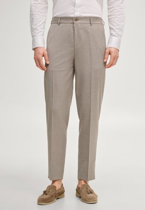 Mężczyzna ubrany w beżowe, dopasowane spodnie, białą koszulę z długim rękawem oraz brązowe zamszowe mokasyny z frędzlami, stojący na prostym, jasnym tle.