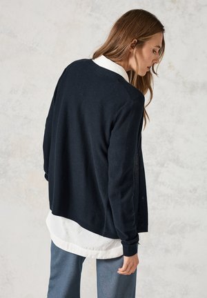 Námořnický pletený cardigan s dlouhými rukávy, otevřeným předním dílem a texturovaným povrchem, vrstvený přes bílou košilovou košili, stylizovaný s světle modrými kalhotami.