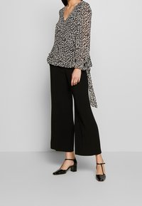 Blouse à motifs noir et blanc avec un col en V, manches longues et poignets à volants, associée à un pantalon noir à jambe large et des talons noirs.