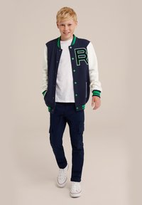 Marinblå varsityjacka med vita ärmar, gröna detaljer och en stor "R"-logotyp. Matchas med marinblå cargobyxor och vita sneakers.