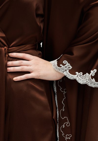 Χέρι που ακουμπά στη μέση, φορά καφέ σατέν ρόμπα δεμένη με ζώνη, η διπλανή ρόμπα έχει άσπρη δαντέλα στο μανίκι.