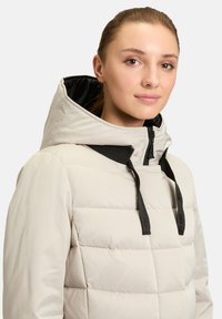 Doudoune beige clair avec un motif matelassé texturé, cordons de serrage noirs réglables et un col haut avec fermeture éclair.