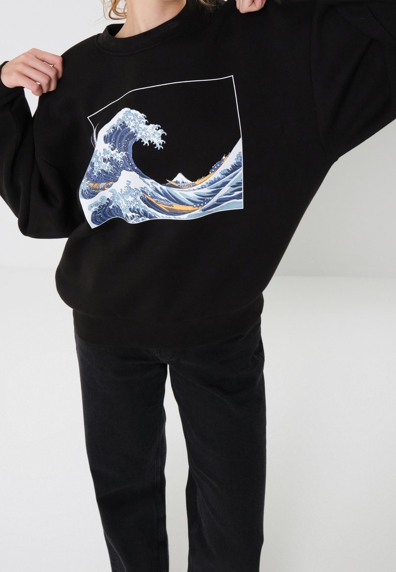 Personne portant un sweat-shirt noir avec une vague océanique bleue et blanche, des bateaux et une montagne en arrière-plan.