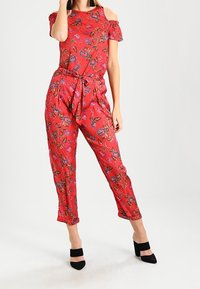 Röd blommig byxdress med off-shoulder ärmar, knytliv, korta ben och svarta högklackade skor. Slät tygtextur.