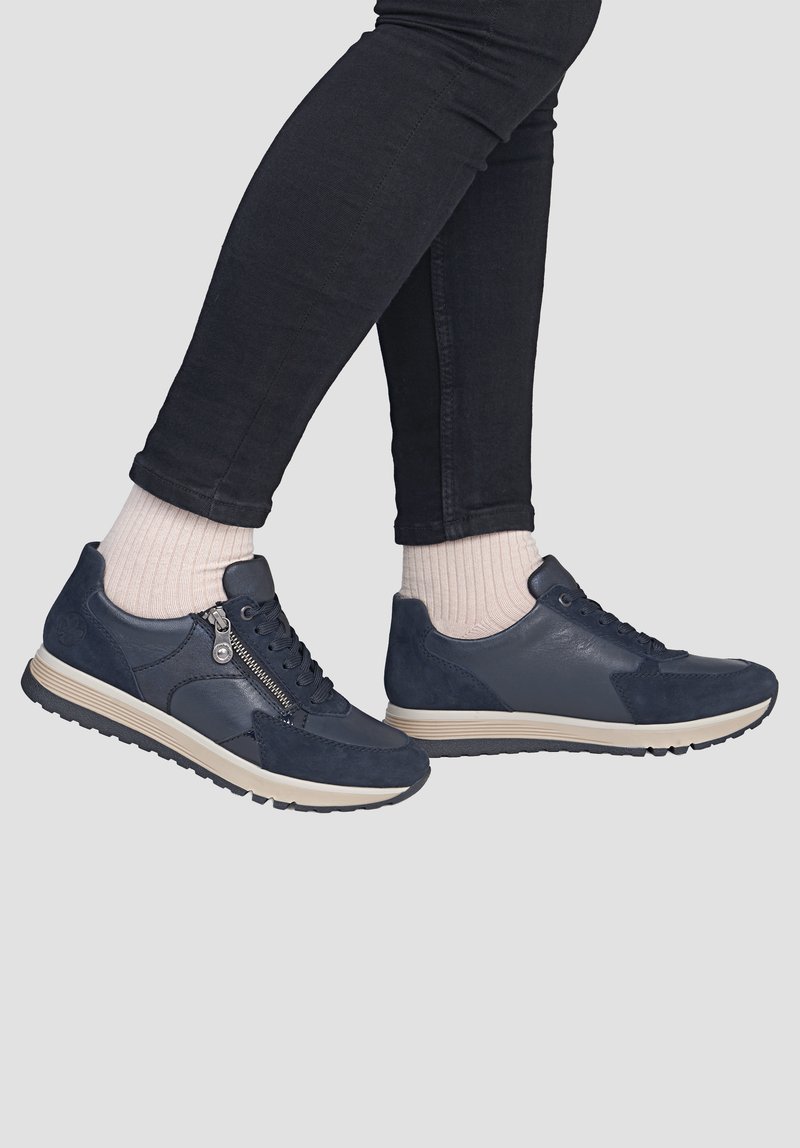 Personne portant des baskets bleu foncé avec des détails zip sur le côté, des chaussettes beiges et un pantalon noir ajusté sur un fond clair uni.