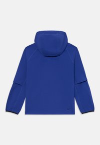 Chaqueta azul con capucha y mangas largas, que presenta una textura suave y un diseño minimalista. Incluye un logo sutil en la parte inferior izquierda.