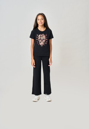 Fille debout portant un t-shirt noir avec un motif de crâne fleuri et de papillon, pantalon noir et baskets blanches sur un fond uni.