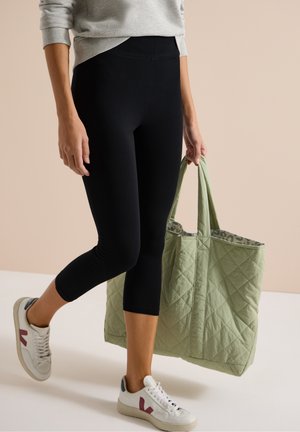 Personne portant des leggings noirs courts, un sweat-shirt gris, des baskets blanches avec des accents roses, et portant un grand sac cabas matelassé vert clair.