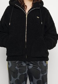 Sudadera con cremallera en forro polar negro, con capucha, bolsillos frontales y cremallera dorada. Presenta un pequeño logo dorado en la parte superior izquierda del pecho. Material texturizado.