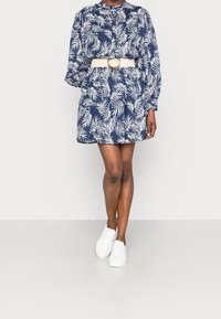 Robe marine avec motif de feuilles blanches, manches longues, devant boutonné, taille cintrée avec une ceinture beige, portée avec des baskets blanches.