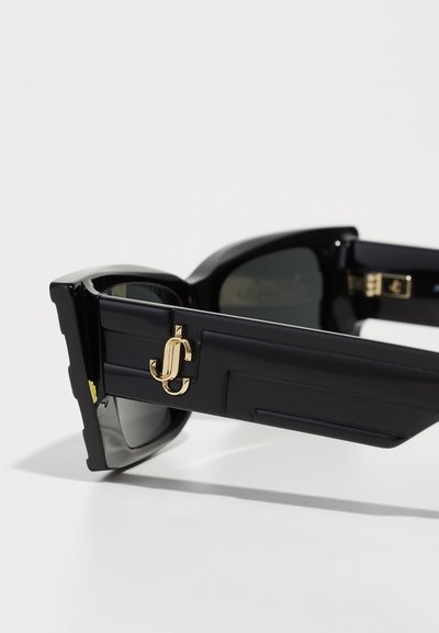 Black sunglasses with dark lenses and a bold rectangular frame. Features a gold accent logo on the side and a textured finish.