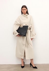 Bež plašč s pasom in zlatimi gumbi, v kombinaciji s črno strukturirano torbico z zlatim poudarkom. Črne špičaste petke.
