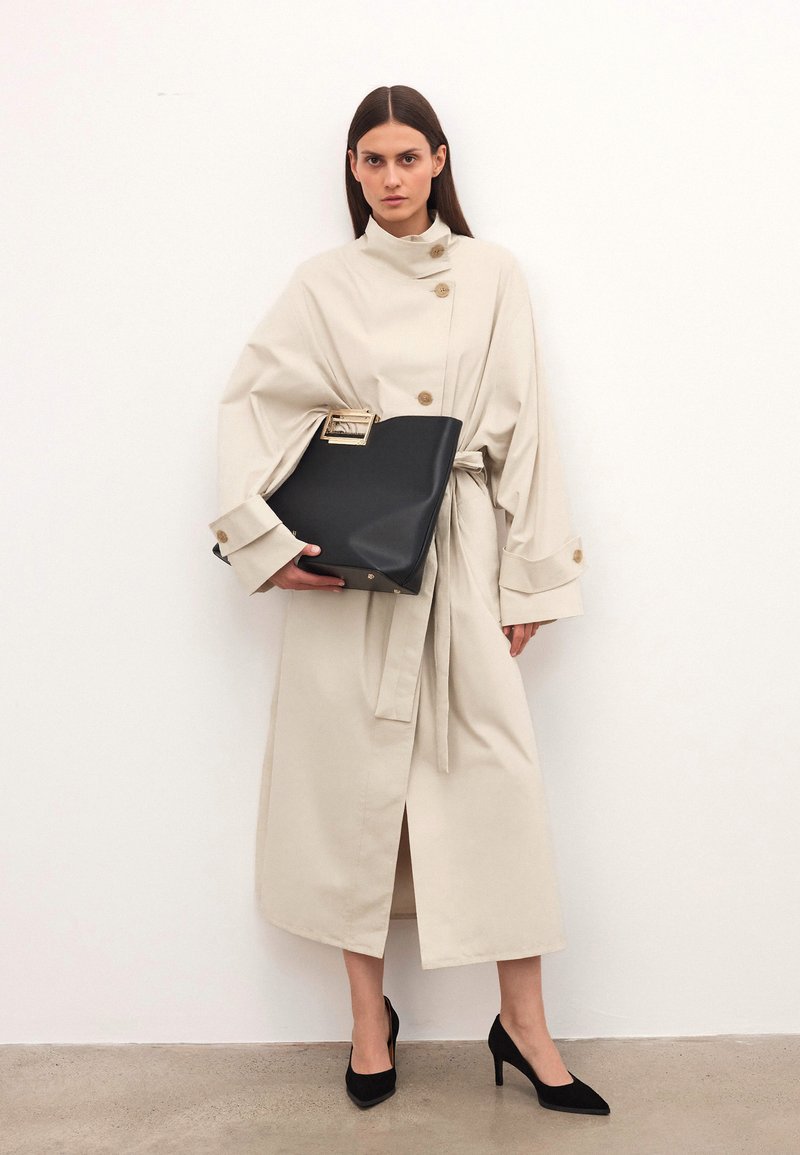 Bež plašč s pasom in zlatimi gumbi, v kombinaciji s črno strukturirano torbico z zlatim poudarkom. Črne špičaste petke.
