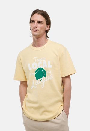 Homme portant un t-shirt jaune pâle avec un graphique de balle de tennis verte et le texte "Tennis Local League 1994", mains dans les poches, regardant légèrement vers la droite.
