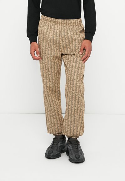 Personne portant un pantalon beige à motifs avec des poignets élastiques, un haut noir à manches longues et des baskets noires, se tenant devant un fond blanc uni.