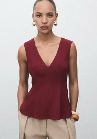 Burgundy peplumtop med djup V-ringning, tillverkad av glatt tyg. Har ärmlös design och subtila sömdetaljer. I kombination med beiga byxor.