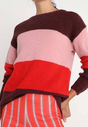 Vrouw draagt een color-block trui met bordeauxrood, roze en rode strepen, gecombineerd met rood-witte broek met verticale strepen.