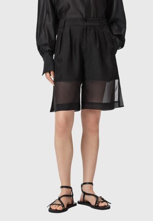 Czarne krótkie spodenki z wysokim stanem i przezroczystymi panelami powyżej kolan, zestawione z czarną bluzką z długim rękawem oraz czarnymi płaskimi sandałami na paski.