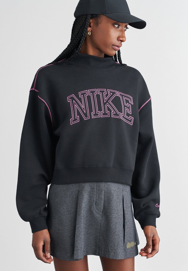 Melna Nike džemperis ar rozā izšūtu logotipu, īsā dizainā un ribinātām aprocēm; kopā ar pelēku plisētu svārku, demonstrējot ikdienišķu sporta stilu.