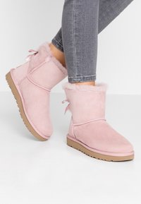 Bottes courtes en daim rose avec doublure en fourrure douce, semelle en caoutchouc plate et un petit nœud à l'arrière. Portées avec un pantalon en jean gris.