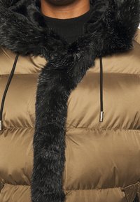 Chaqueta de plumas marrón con capucha forrada de piel sintética negra, cuello acanalado y detalles de cordón. Resistente al agua y con textura acolchada.