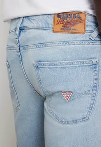 Jeans de mezclilla azul claro con un acabado texturizado, que presentan un parche con un logo en forma de triángulo en el bolsillo trasero y una etiqueta de cuero marrón en la cinturilla.