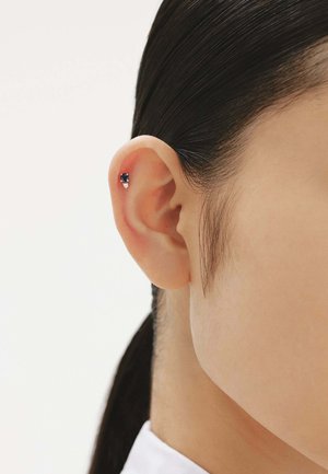 Pendiente pequeño de stud con una gema azul oscura y acentos plateados, situado en la parte superior de la oreja. Textura de piel suave visible.