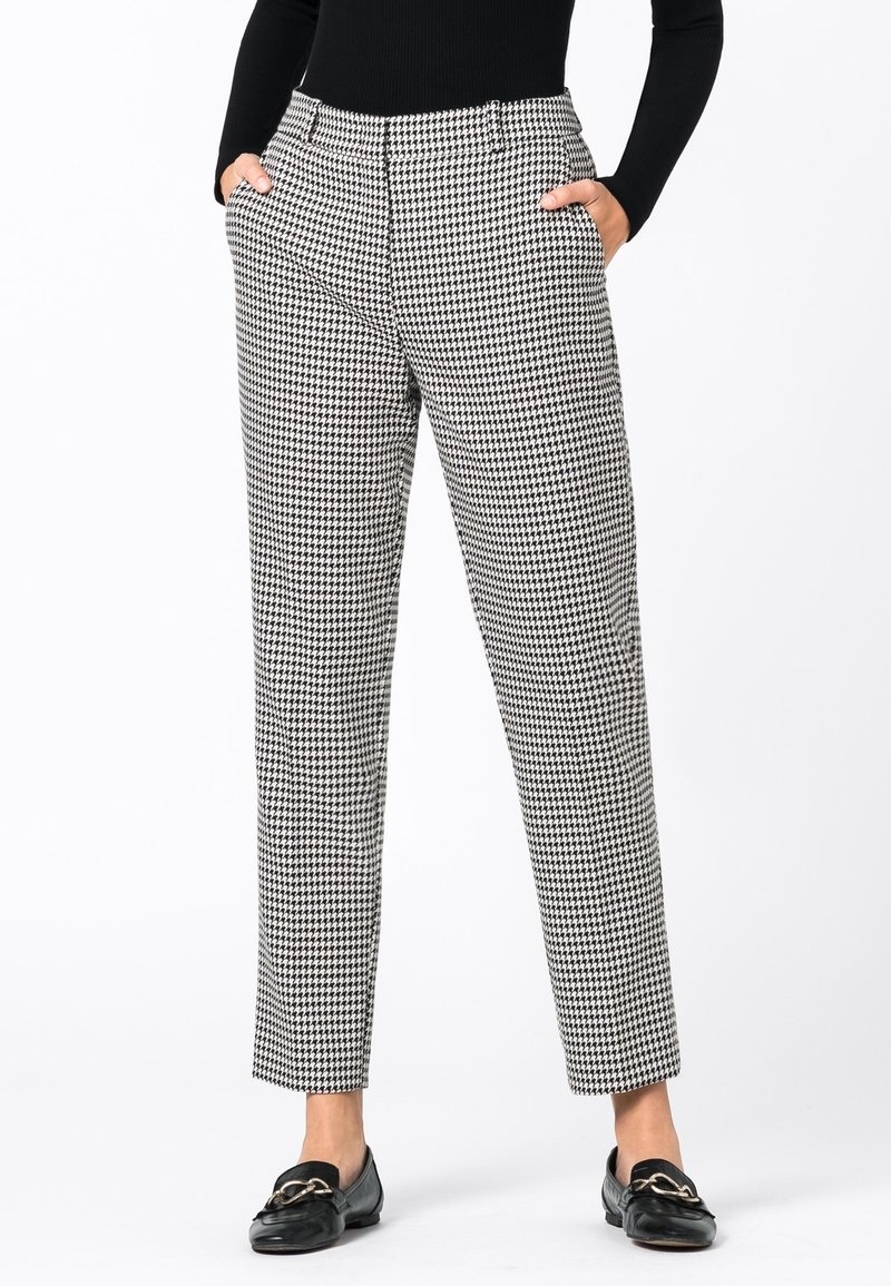 Femme portant un pantalon pied-de-poule noir et blanc, un top à manches longues noir, les mains dans les poches, et des mocassins noirs avec un détail de chaîne dorée.