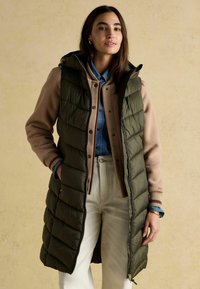 Kobieta ubrana w długi oliwkowo-zielony pikowany kamizelkę na beżowej kurtce, niebieskiej koszuli dżinsowej i białych spodniach, stojąca z jedną ręką w kieszeni.