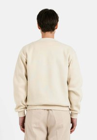 Sweatshirt beige con cuello redondo y puños acanalados. Textura suave, corte relajado y diseño minimalista en la parte posterior. Combinado con pantalones beiges.
