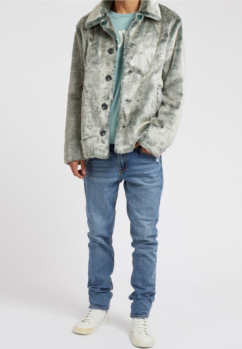 Veste en fausse fourrure vert clair avec un col et des boutons noirs, portée sur une chemise bleu clair, associée à un jean bleu et des chaussures blanches.