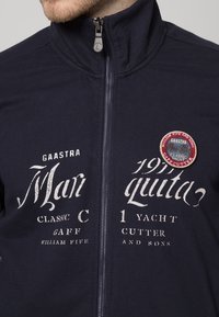 Marinblå zip-jacka i bomullsmix, prydd med vit tryckt text och ett runt tygmärke med maritim design. Slät yta, hög krage.