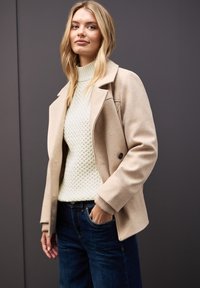 Beige ullblandad jacka med spetsig krage, framknappar och sidfickor, över en ljusgrön texturerad tröja och mörkblå jeans.