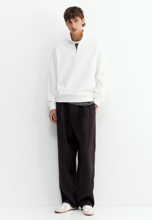 Jeune homme debout contre un mur blanc portant un sweat-shirt blanc à fermeture éclair quart de zip, un pantalon large noir et des baskets blanches avec des semelles claires.