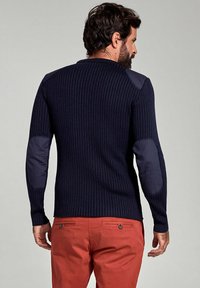 Maglione navy a costine con toppe blu scuro sui gomiti, scollo rotondo e maniche lunghe, abbinato a pantaloni rossi, visto da dietro.