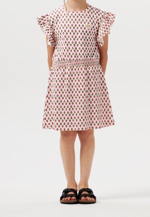 Niña con vestido blanco estampado con motivos florales en rosa y marrón, mangas con volantes y sandalias negras con hebilla, de pie sobre un suelo blanco liso.