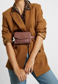 Blazer marrón sobre una camisa de estampado animal, con un bolso de cintura de piel malva que presenta un cierre de solapa y costuras blancas, llevado alrededor de la cintura.